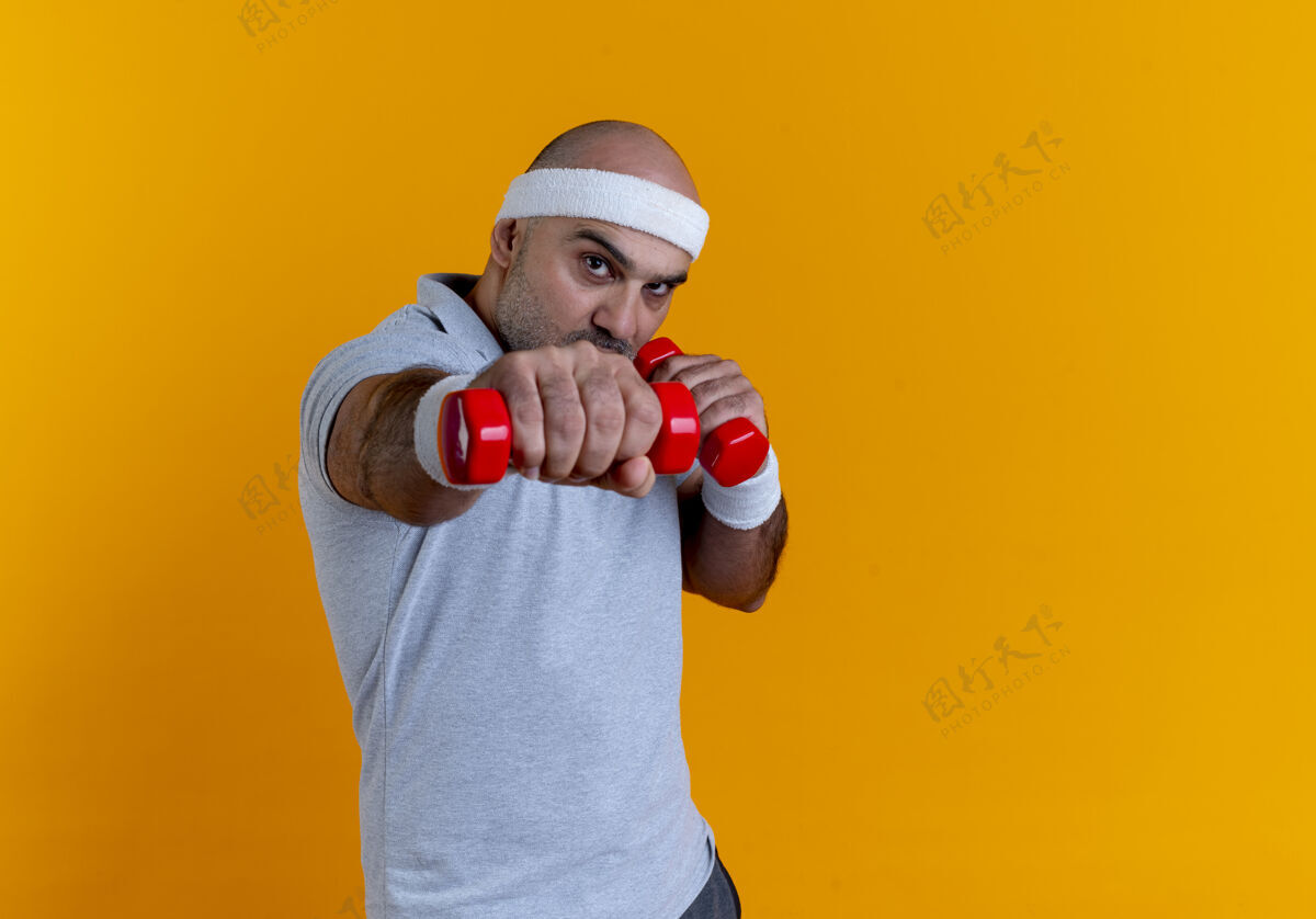 看戴着头巾的成熟运动型男人 站在橙色的墙上 带着看起来紧张而自信的哑铃锻炼运动员男人男人