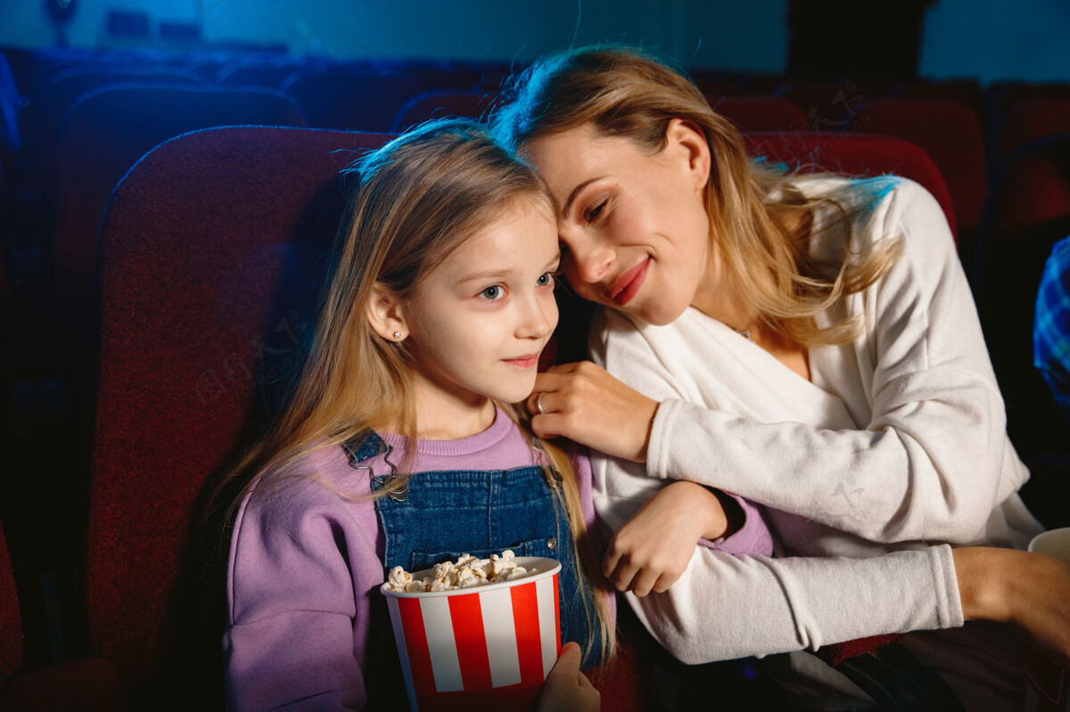 幽默白种人的母亲和女儿在电影院 房子或电影院看电影看起来富有表现力 惊讶和情绪化独自坐着玩关系 爱情 家庭 童年 周末娱乐电影房子