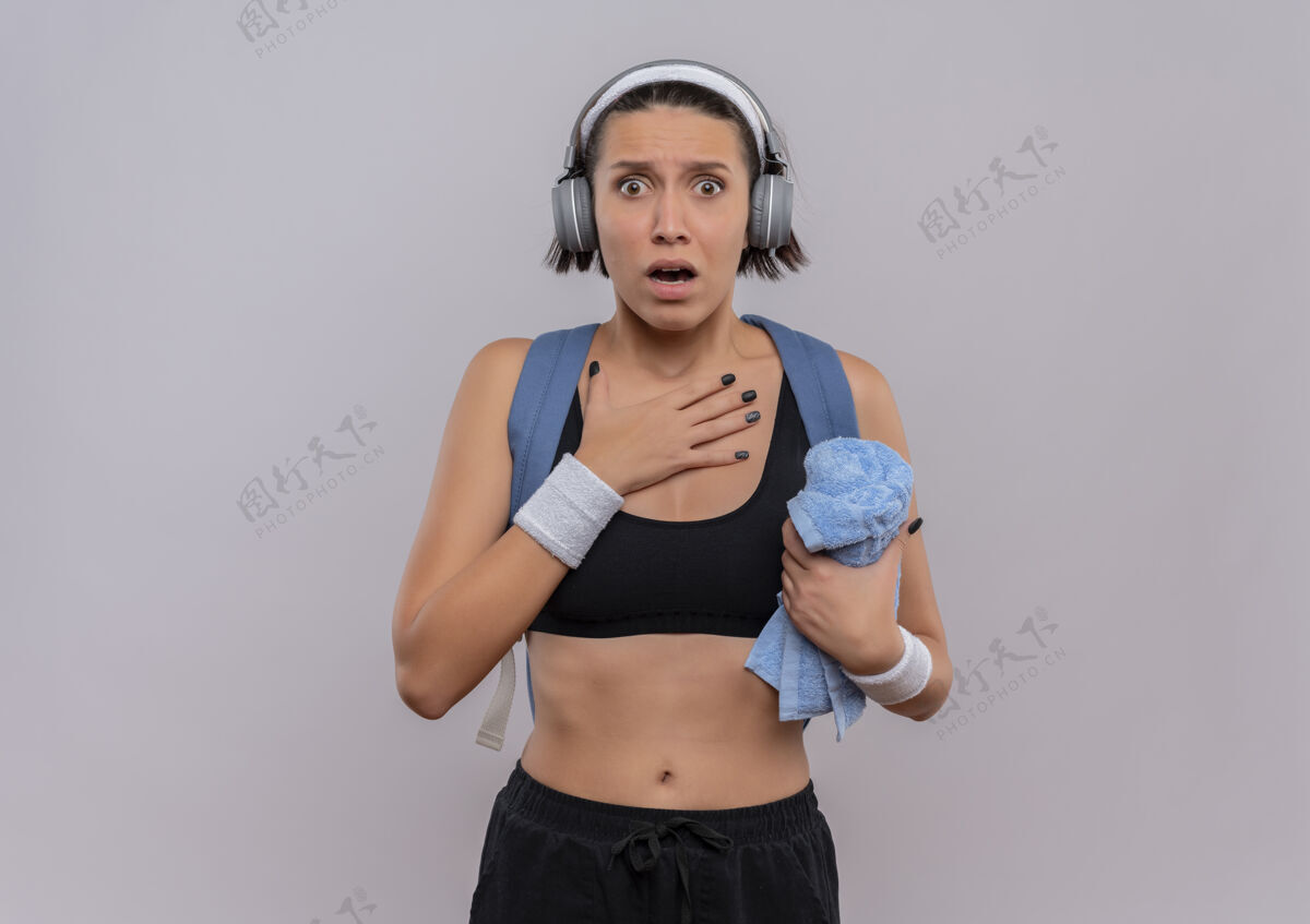 耳机年轻的健身女士穿着运动服 背着背包 头上戴着耳机 手放在胸前拿着毛巾 站在白墙上吓了一跳胸前抱着手