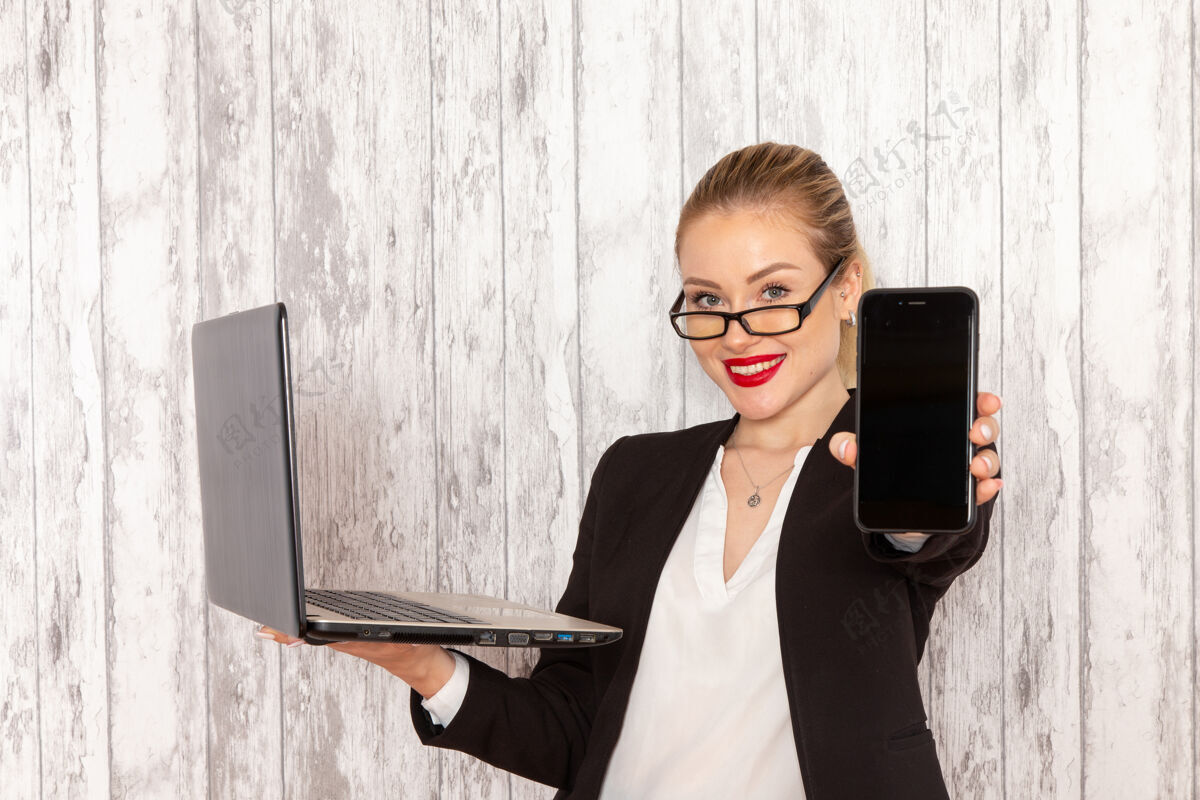 衣服正面图：穿着严苛衣服的年轻女商人 黑色夹克 手持手提电脑 在白色的表面上微笑着正面笔记本电脑电话