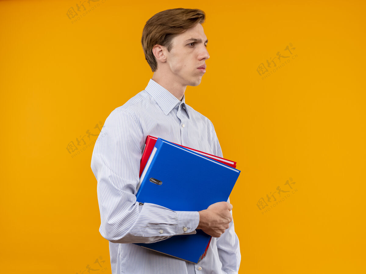 严肃一个穿着白衬衫的年轻人拿着文件夹 严肃地站在橙色的背景上 脸上带着不高兴的表情 一边看着衬衫夹着不高兴