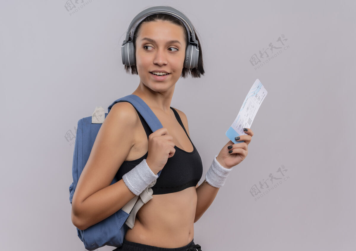 运动员身穿运动服 头戴耳机 手持机票 面带微笑地站在白墙上的年轻健身女士头看健身
