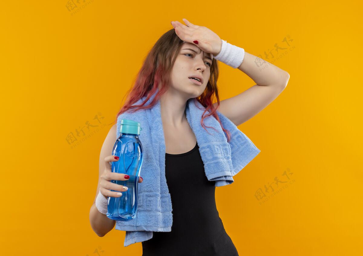 健身年轻的健身女孩穿着运动服 脖子上挂着毛巾 手里拿着一瓶看起来不舒服的水 站在橘色的墙上摸着她的头触摸人站