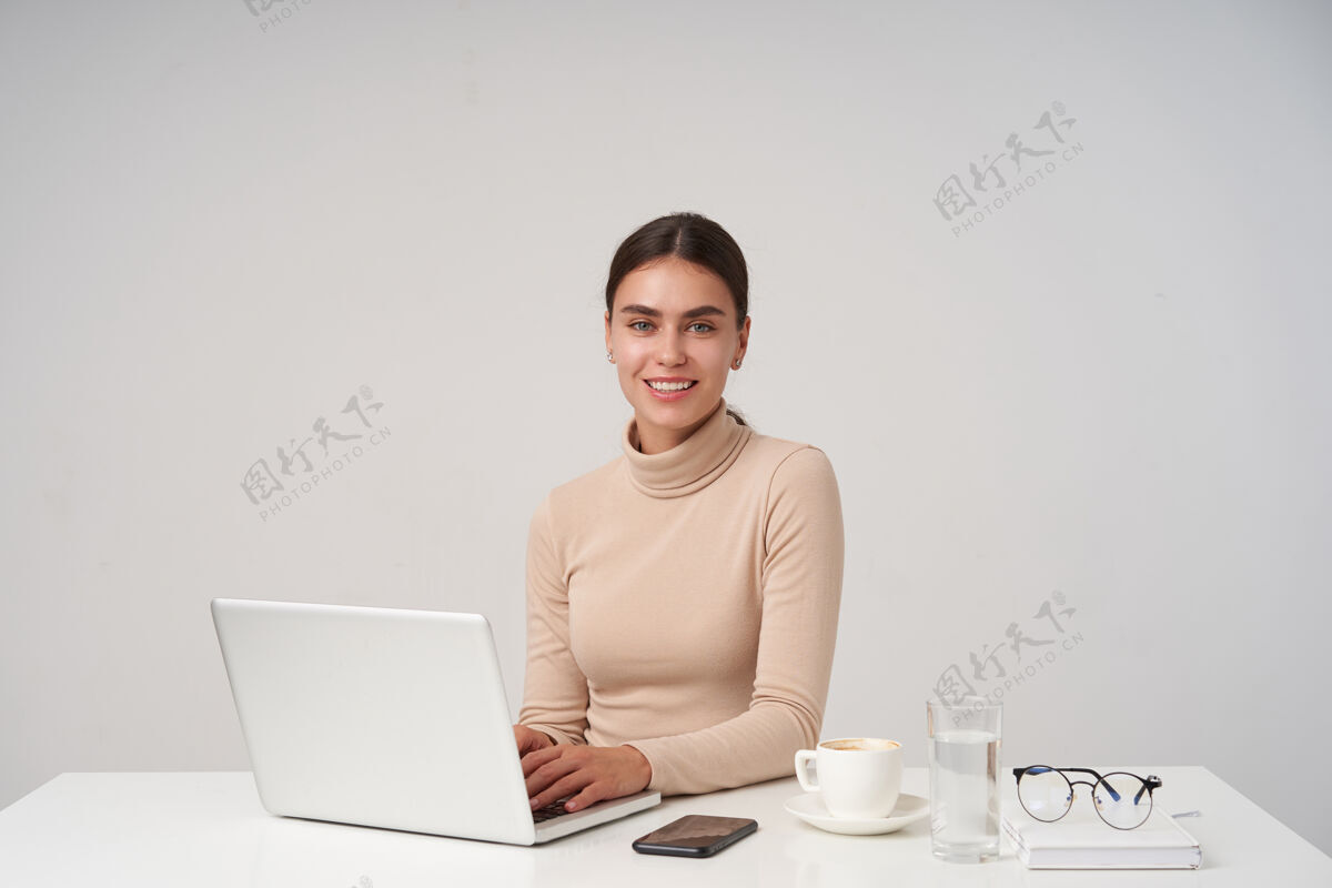 玻璃一张室内照片 欢快的年轻漂亮的黑发女性露出她完美的白牙 面带微笑 一边在键盘上打字 一边在白墙上摆姿势Mua正式头发