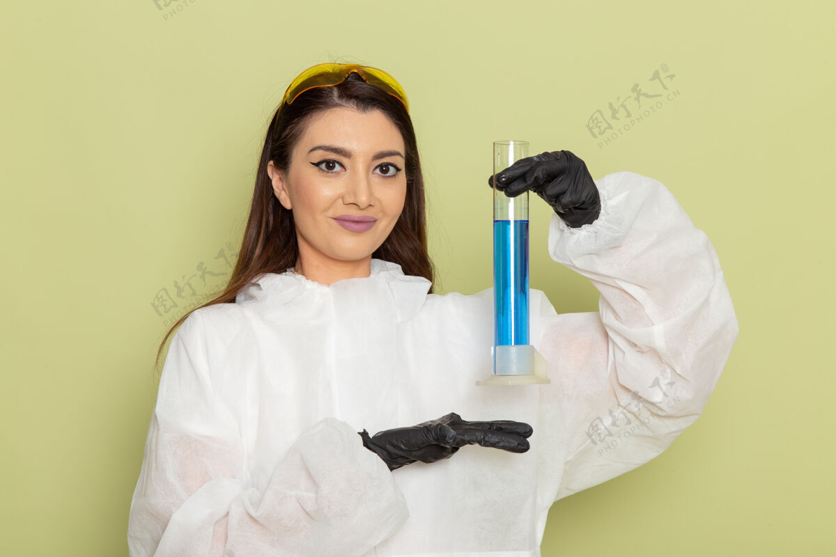 化学家正面图穿着特殊防护服的女化学家拿着装有蓝色溶液的烧瓶 在绿色表面微笑女化学品正面