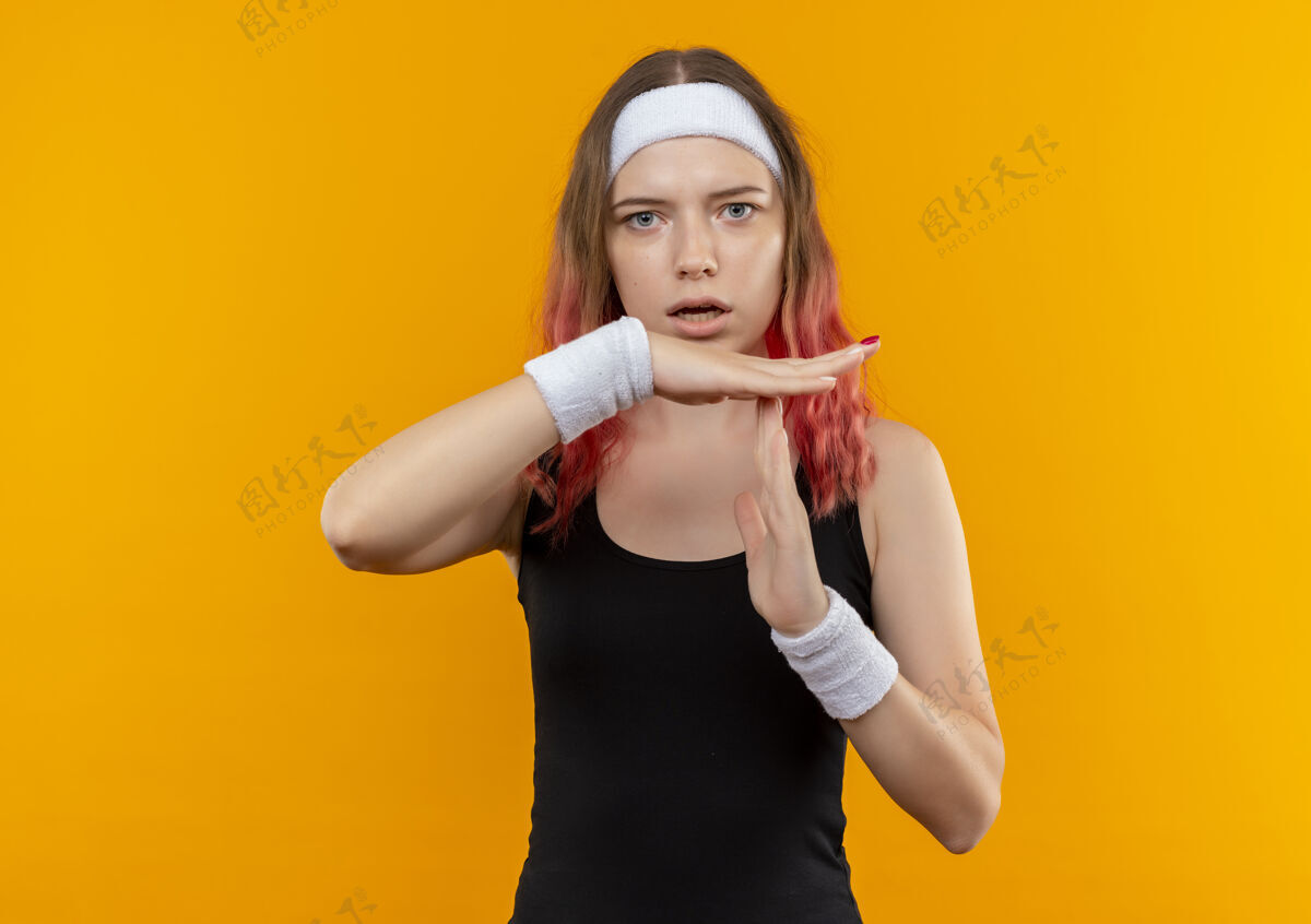 年轻穿着运动服的年轻健身女士站在橙色的墙上做着暂停的手势人时间运动