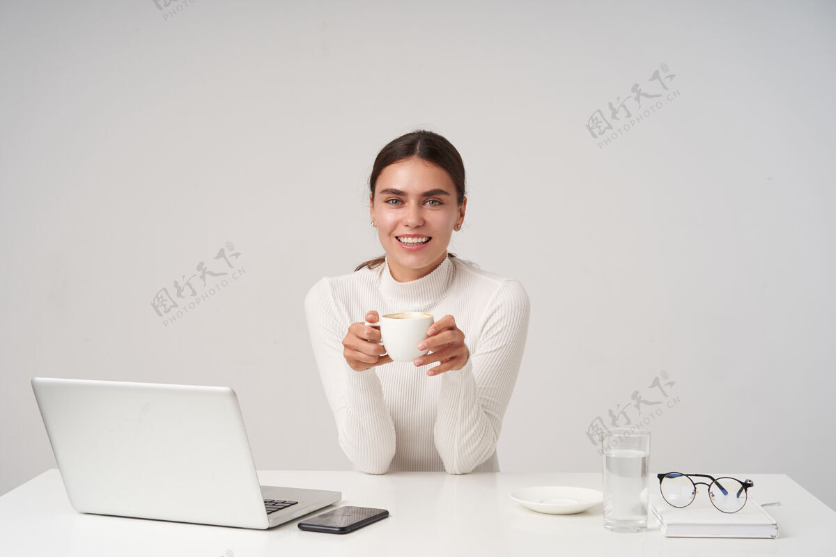 优雅照片中 年轻迷人的黑发女子穿着白色针织马球衫 坐在桌旁 手举茶杯 面带微笑 神情欢快 隔着白色的墙壁微笑女士正式