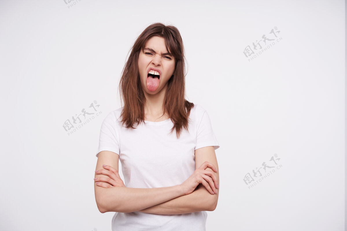 休闲这张照片拍摄的是一位兴奋的年轻黑发女性 随意的发型双手交叉放在胸前 露出舌头 穿着基本款t恤在白墙上摆姿势砰额头20岁