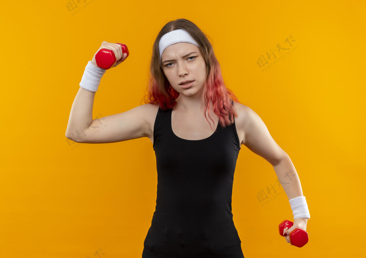 运动装穿着运动服的年轻健身女士手拿哑铃 站在橘色的墙上做运动 看起来很累手运动员看