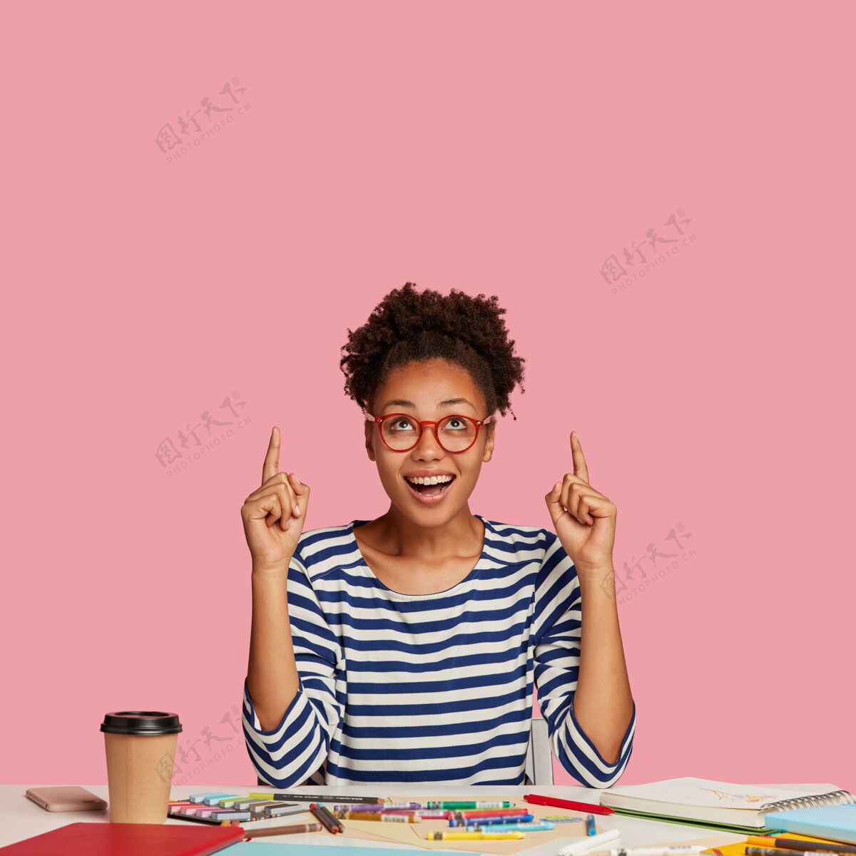 写作快乐黑皮肤的可爱女艺术家的垂直镜头 用食指指着 穿着条纹毛衣 模特对着粉色的墙壁 感觉很兴奋 用螺旋笔记本摆在桌面上钢笔喝酒建筑师