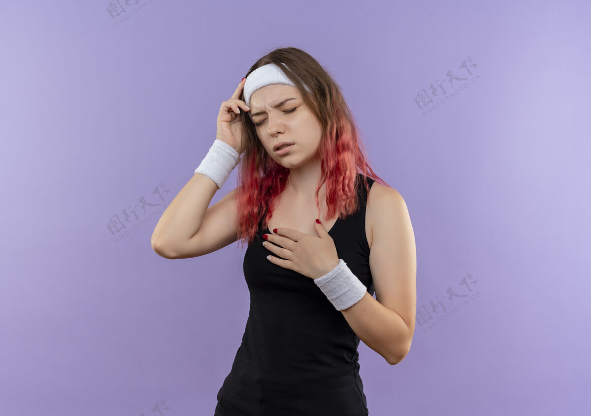 头部穿着运动服的年轻健身女士站在紫色的墙上 看上去身体不适 摸着头痛的头看不适运动员