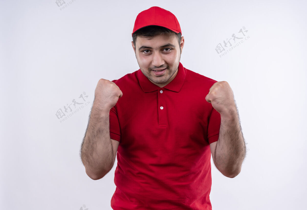帽子身穿红色制服 戴着帽子 握紧拳头的年轻送货员站在白色的墙上 既高兴又兴奋握紧公民快乐