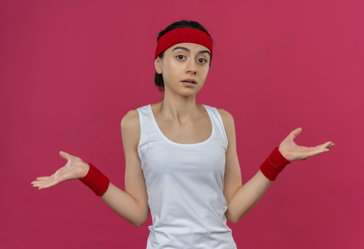 运动身穿运动服的年轻健身女 头上戴着头巾 神情迷茫 不确定地向两边伸开双臂 站在粉色的墙上 没有人回答运动装困惑问答