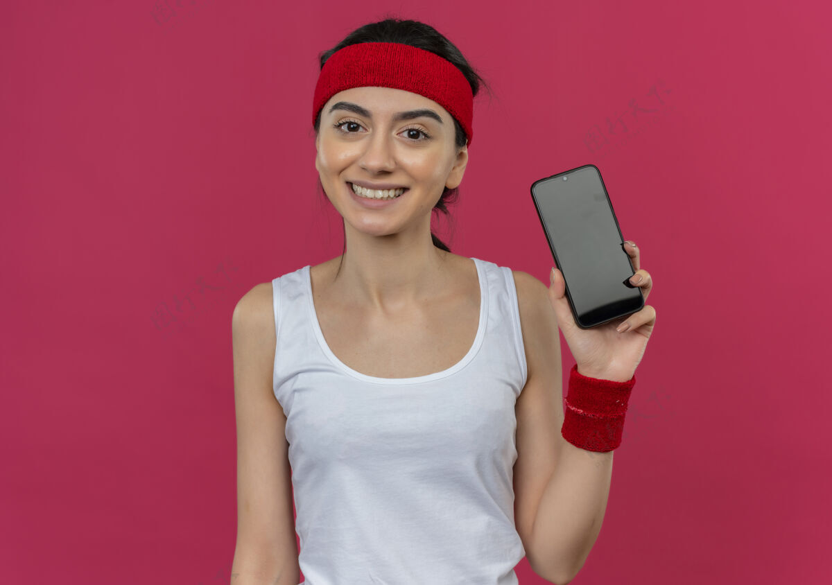 年轻身穿运动服 头戴智能手机的年轻健身女士站在粉红色的墙上 脸上露出快乐和积极的微笑运动人快乐