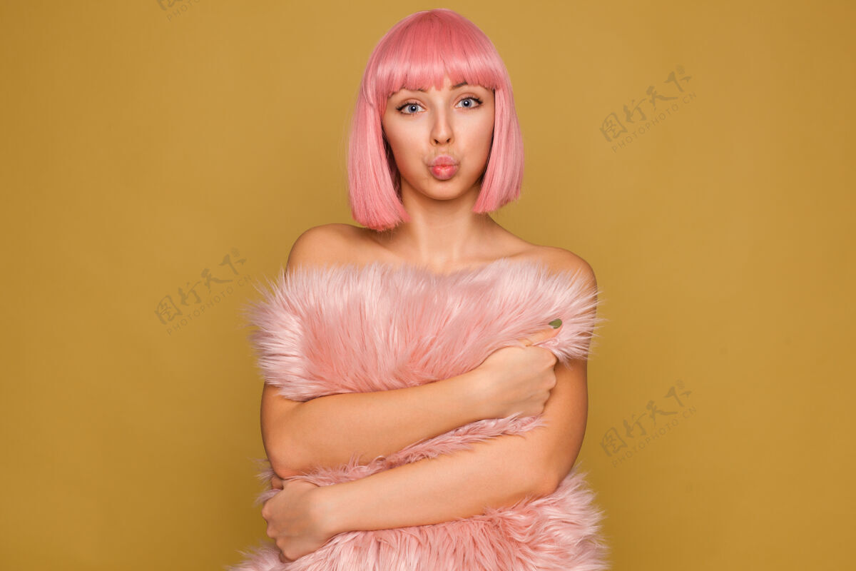 女性室内照片：年轻漂亮的蓝眼睛粉红头发女士撅着嘴 兴奋地看着 手里拿着蓬松的枕头 站在芥末墙上年轻邦短裤