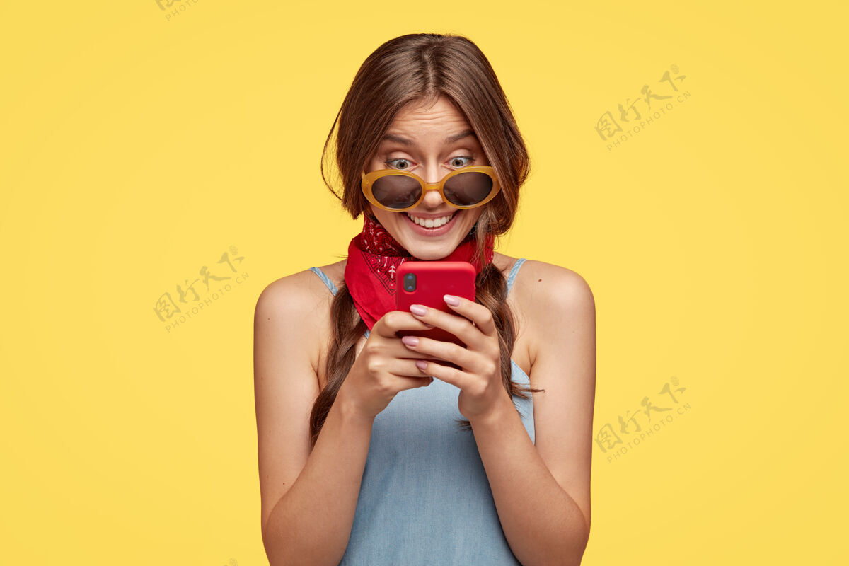 牙关笑容可掬的黑发女子表情欢快 手持红色手机 乐于阅读短信 连接无线互联网 隔着黄墙与世隔绝人 科技 休闲女朋友女人小工具