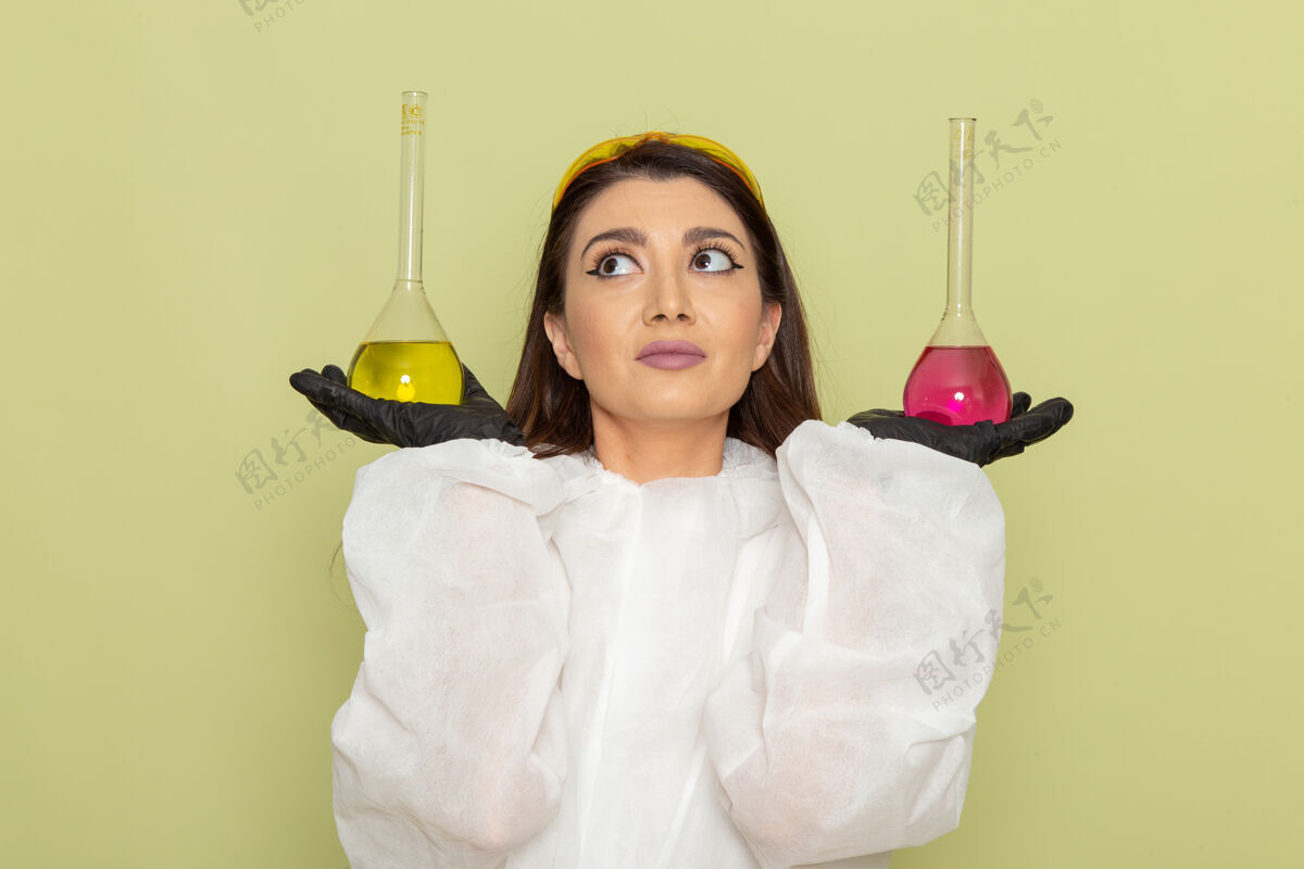 科学前视图穿着特殊防护服的女化学家 手拿淡绿色表面上的粉色和黄色溶液化学化学漂亮