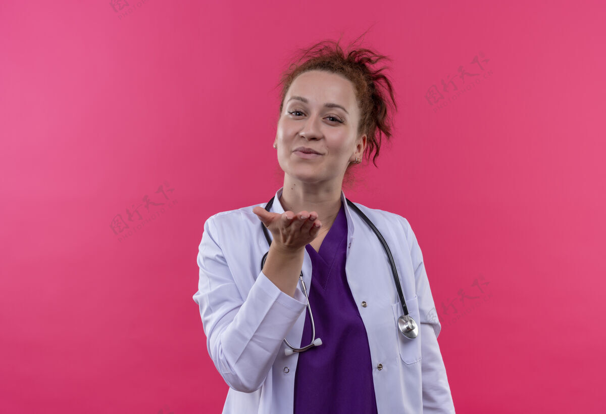 亲吻年轻的女医生穿着白大褂 手持听诊器 站在粉红色的墙上 用手在空气中吹着吻空气外套爱