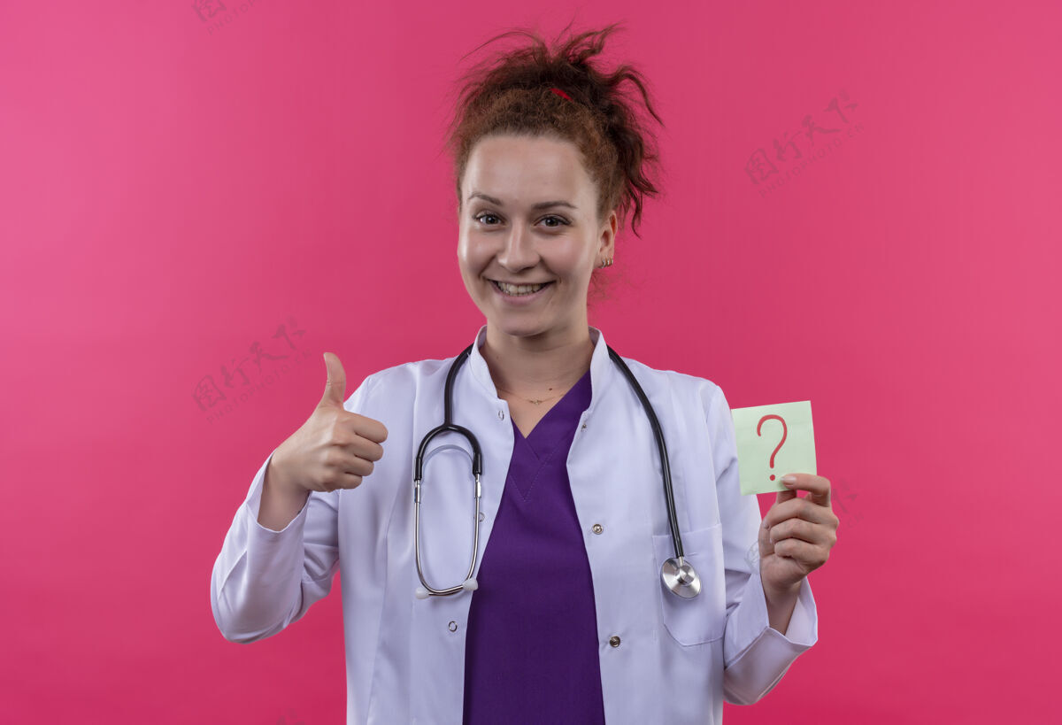 医生年轻的女医生身穿白色外套 手持听诊器 拿着问号纸微笑着竖起大拇指站在粉色的墙上听诊器马克外套