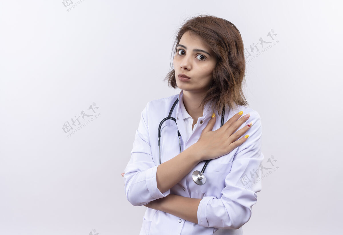 医生身穿白大褂 手持听诊器的年轻女医生站在白墙上 一副忧心忡忡的样子 手放在胸口女人年轻人听诊器