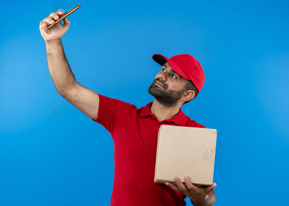 自拍有胡子的送货员穿着红色制服 拿着帽子的盒子 站在蓝色的墙上用他的智能手机自拍站着送货手机
