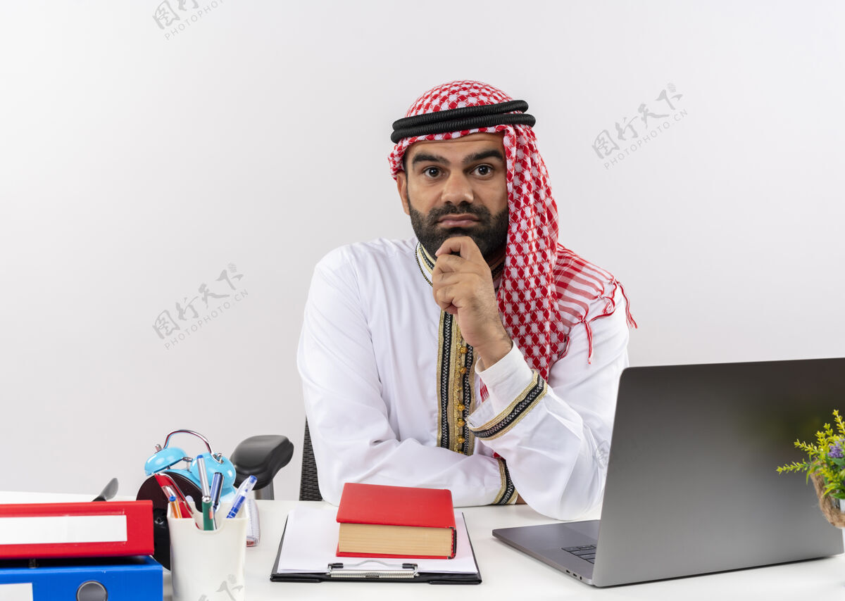 阿拉伯语身着传统服装的阿拉伯商人坐在桌旁 手放在下巴上 困惑地思考着在办公室工作手工作穿