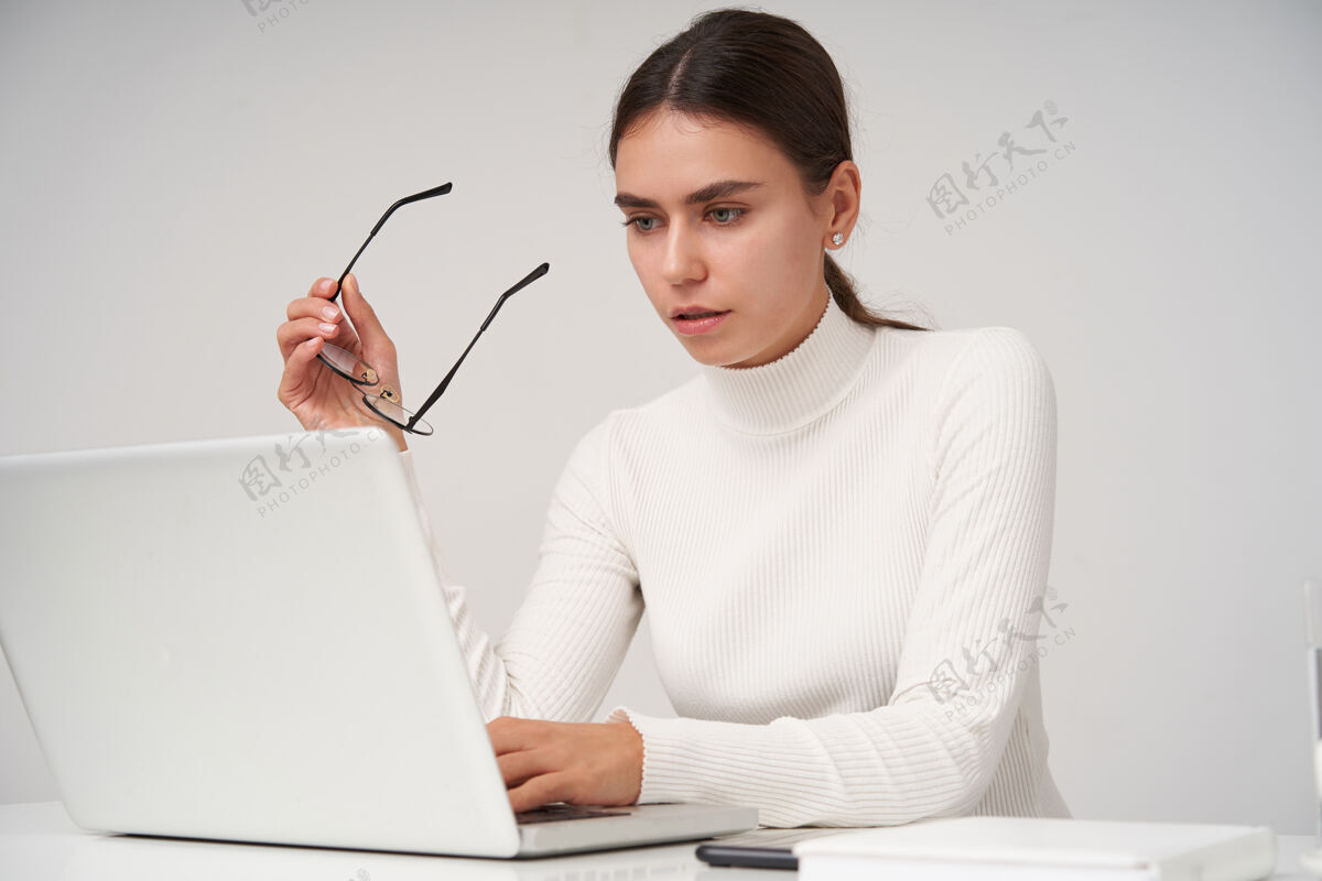 化妆品年轻漂亮的黑发女子 梳着马尾辫 举着眼镜 坐在白色的墙上 凝神地看着她的笔记本电脑专注欧洲穿着