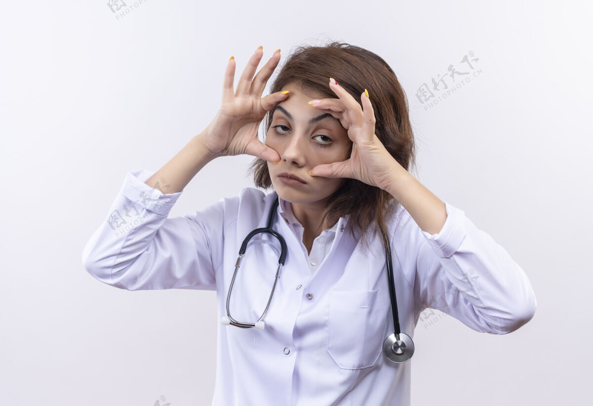 女人身穿白大褂 手持听诊器的年轻女医生站在白墙上试图睁开眼睛 双手感到晨曦疲惫听诊器开试试