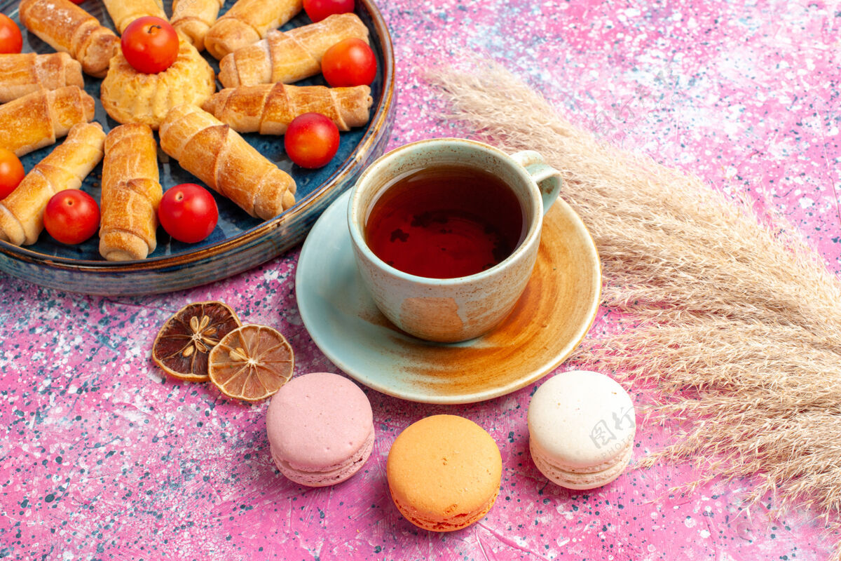 甜点前视图甜甜可口的百吉饼内托盘与新鲜的酸李子法国马卡龙和一杯茶在淡粉色的桌子上派饼干咖啡