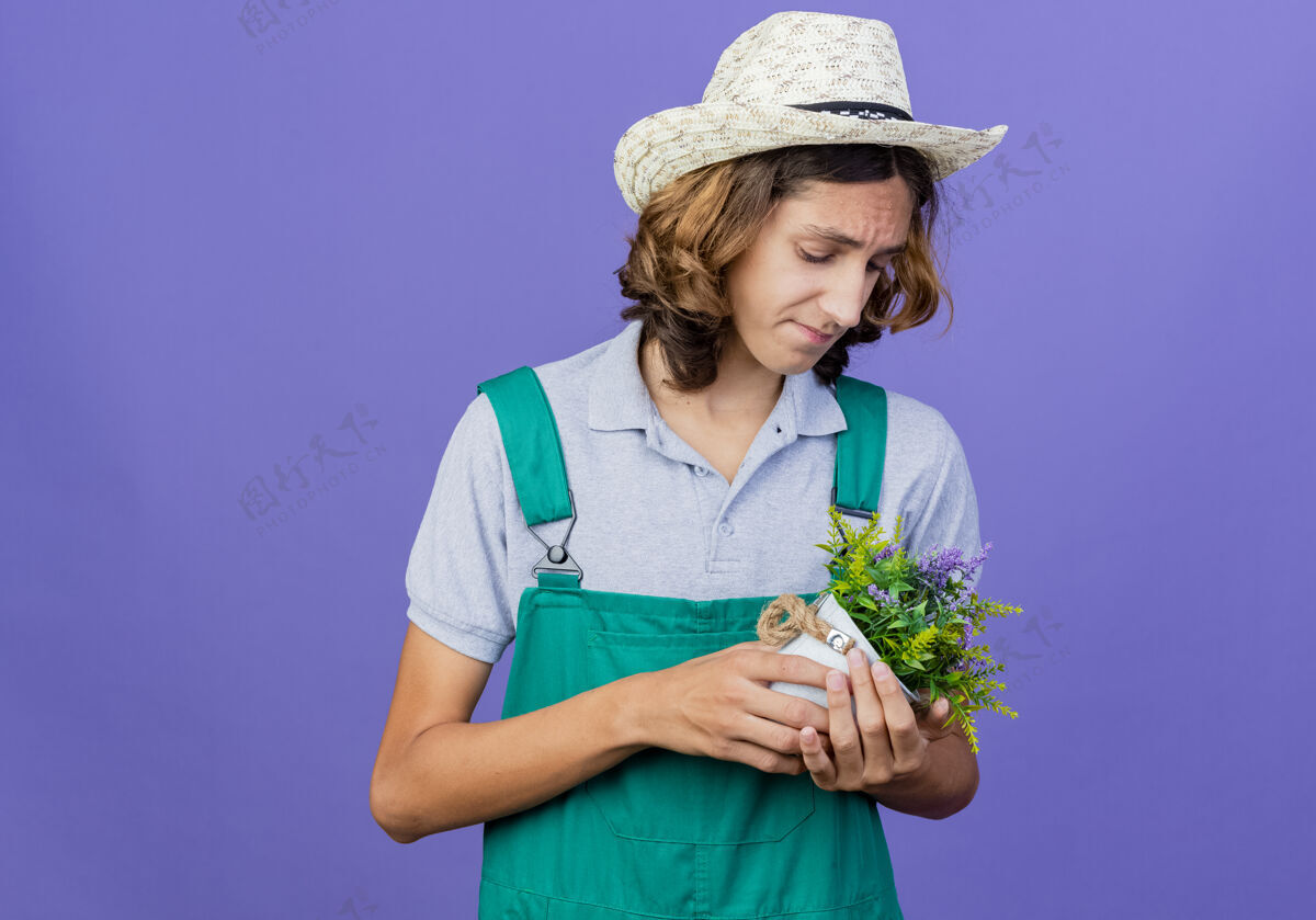 锅年轻的园丁穿着连体衣戴着帽子拿着盆栽植物表情抱着站着