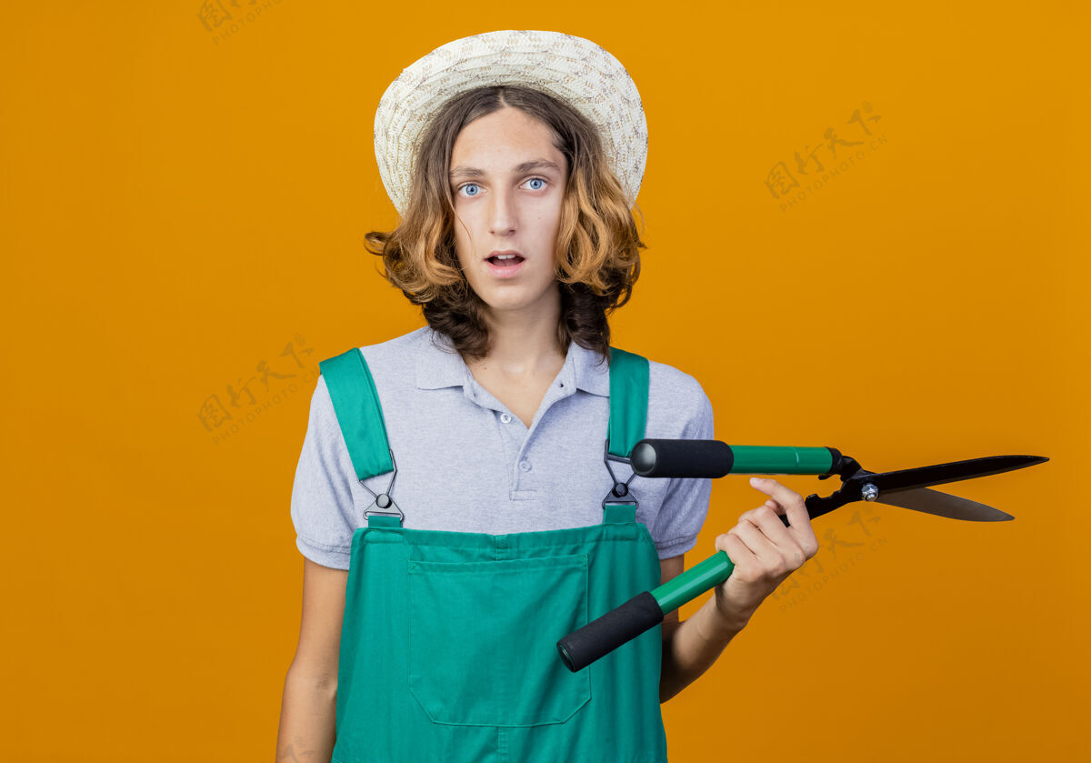 困惑年轻的园丁穿着连体衣戴着帽子拿着树篱剪被搞糊涂了篱笆年轻抱