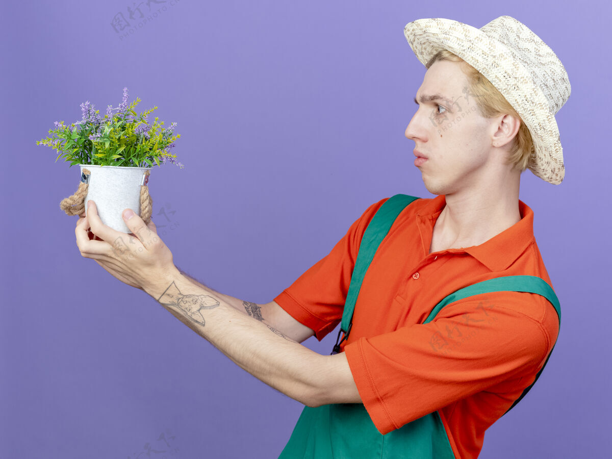 男人年轻的园丁穿着连体衣 戴着帽子 手里拿着盆栽植物 困惑地看着它帽子罐子困惑
