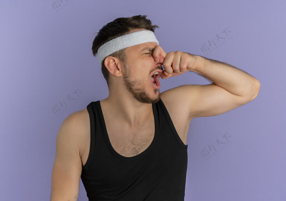 运动员戴着头巾的年轻健身男子站在紫色的墙上用手指捂住鼻子忍受着恶臭封闭男性站