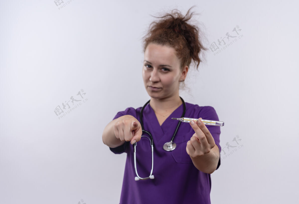 医生年轻的女医生穿着医疗制服 手持听诊器 手指指着温度计 严肃的脸站在白色的墙上年轻人指着脸