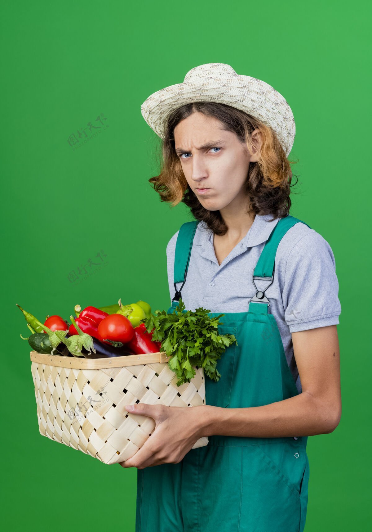 立场年轻的园丁穿着连体衣戴着帽子拿着装满新鲜蔬菜的箱子帽子新鲜板条箱