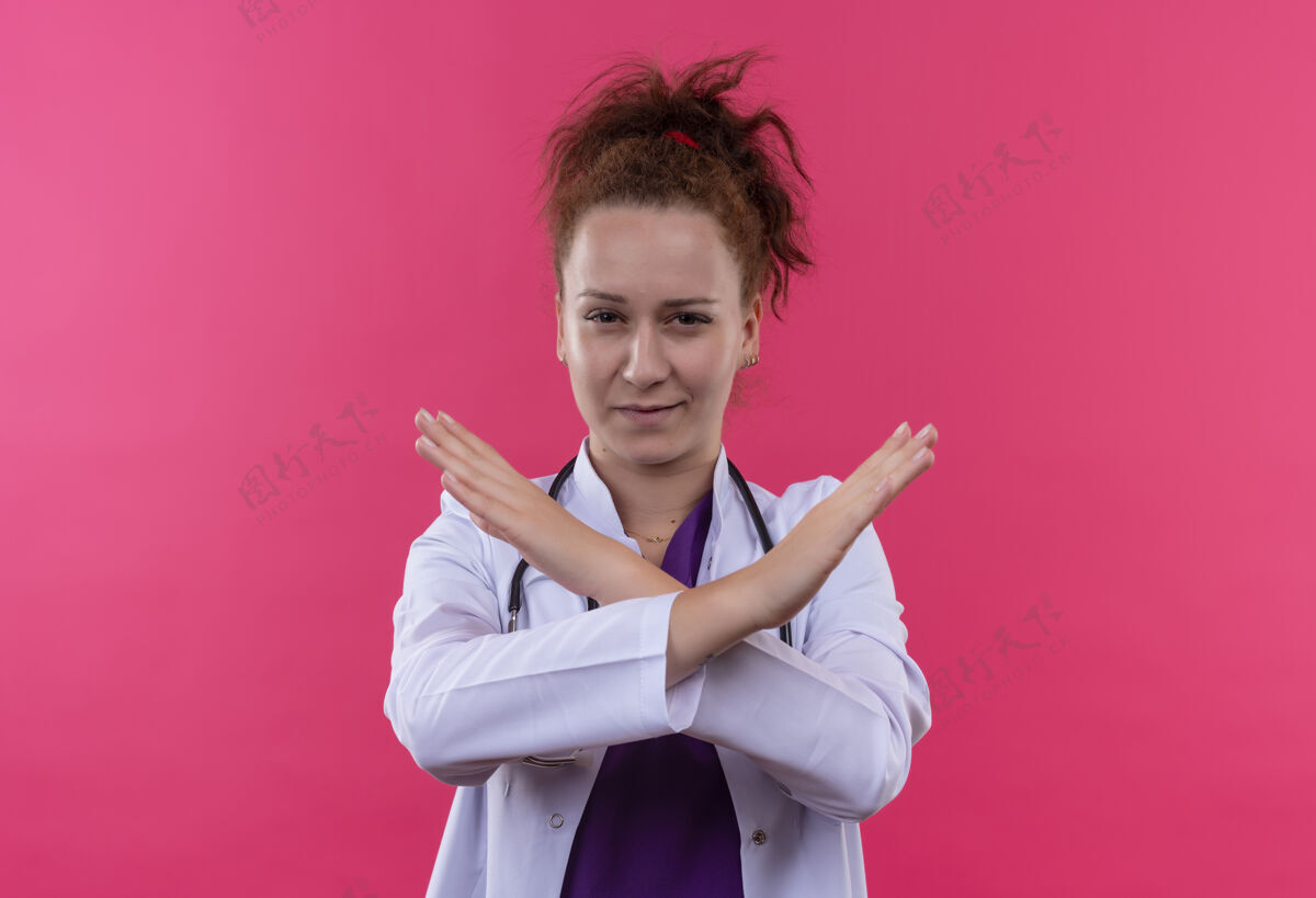 手势年轻的女医生穿着白大褂 手持听诊器 双手交叉 表情严肃地站在粉红色的墙上听诊器医生制造