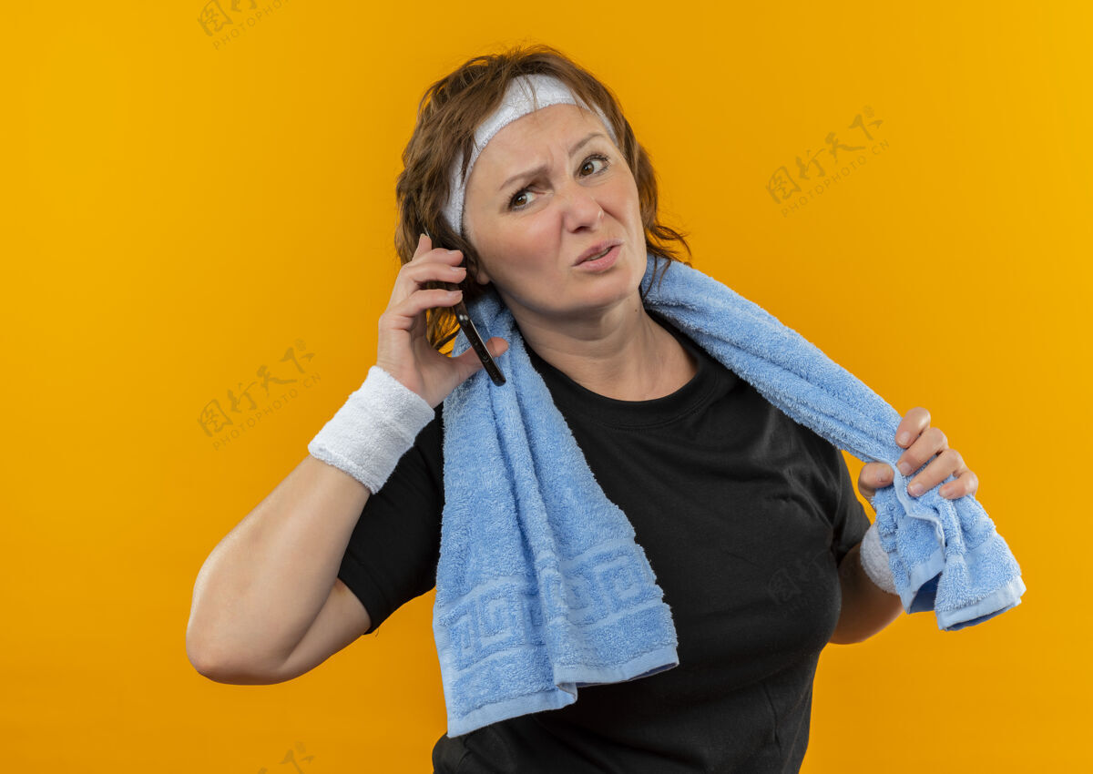 电话中年运动女性 身穿黑色t恤 头上系着头带 肩上搭着毛巾 站在橘色的墙上 看着疲惫地讲着手机说话累肩膀