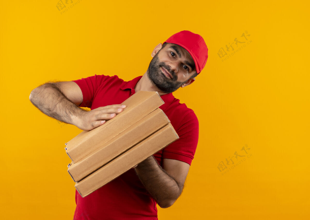 帽子满脸胡须 身穿红色制服 头戴鸭舌帽的送货员站在橙色的墙上 手里拿着一叠披萨盒 脸上带着微笑看送货站着