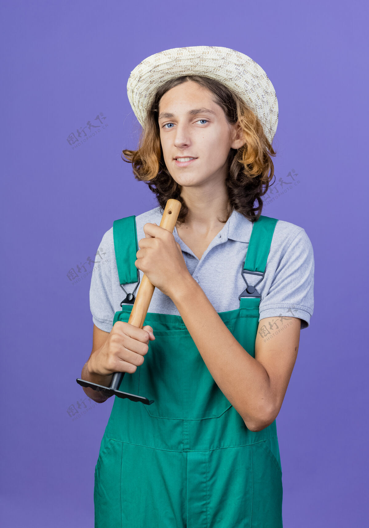 连身衣年轻的园丁 穿着连体衣 戴着帽子 手里拿着迷你耙子 脸上带着微笑抱着帽子花园