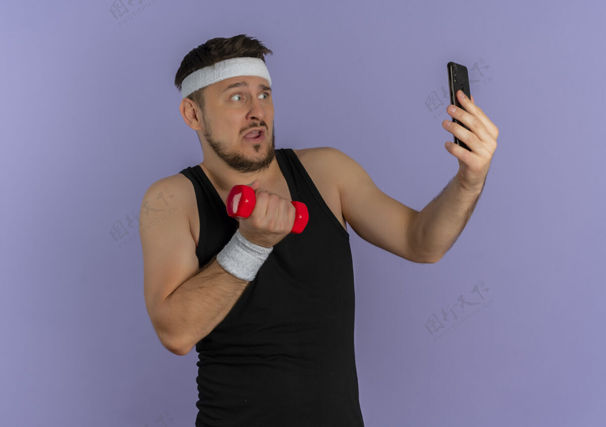 运动员戴着头巾的年轻健身男子用智能手机自拍 手里拿着哑铃 站在紫色的墙上 看上去很困惑采取手移动