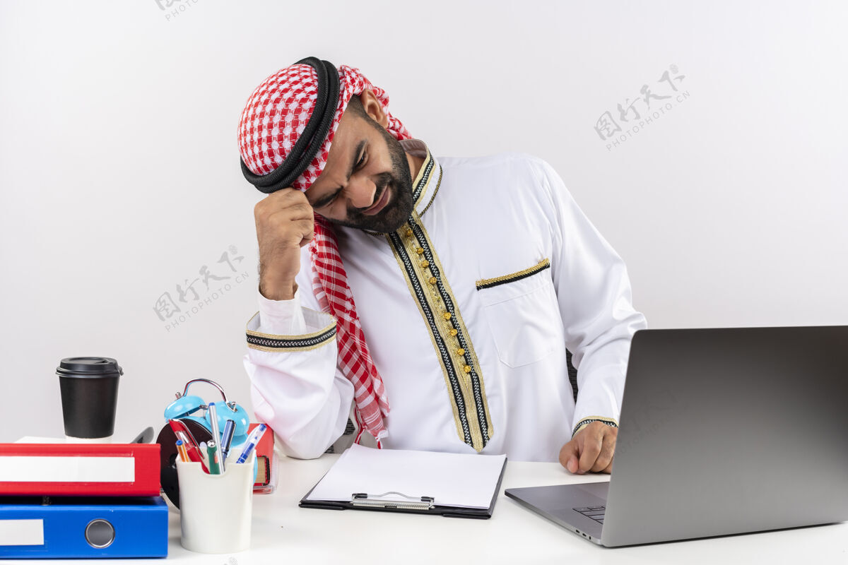 失望身着传统服装的阿拉伯商人坐在桌旁 手提电脑看起来很疲惫 在办公室工作很失望穿阿拉伯语坐