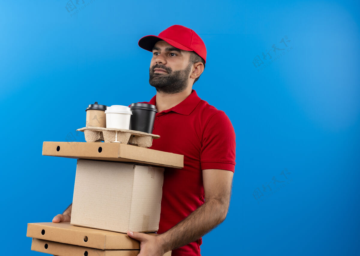 男人一个留着胡须的送货员 穿着红色制服 戴着帽子 手里拿着纸板箱 脸上带着微笑站在蓝色的墙上望着一边站着帽子拿着
