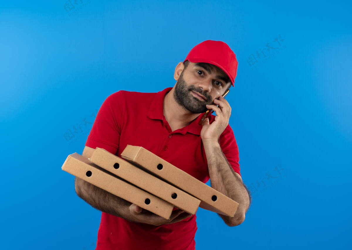 站着站在蓝色的墙上 留着胡须 身穿红色制服 头戴鸭舌帽 手里拿着一叠比萨饼盒 一边微笑一边讲着手机的送货员谈话手机叠着