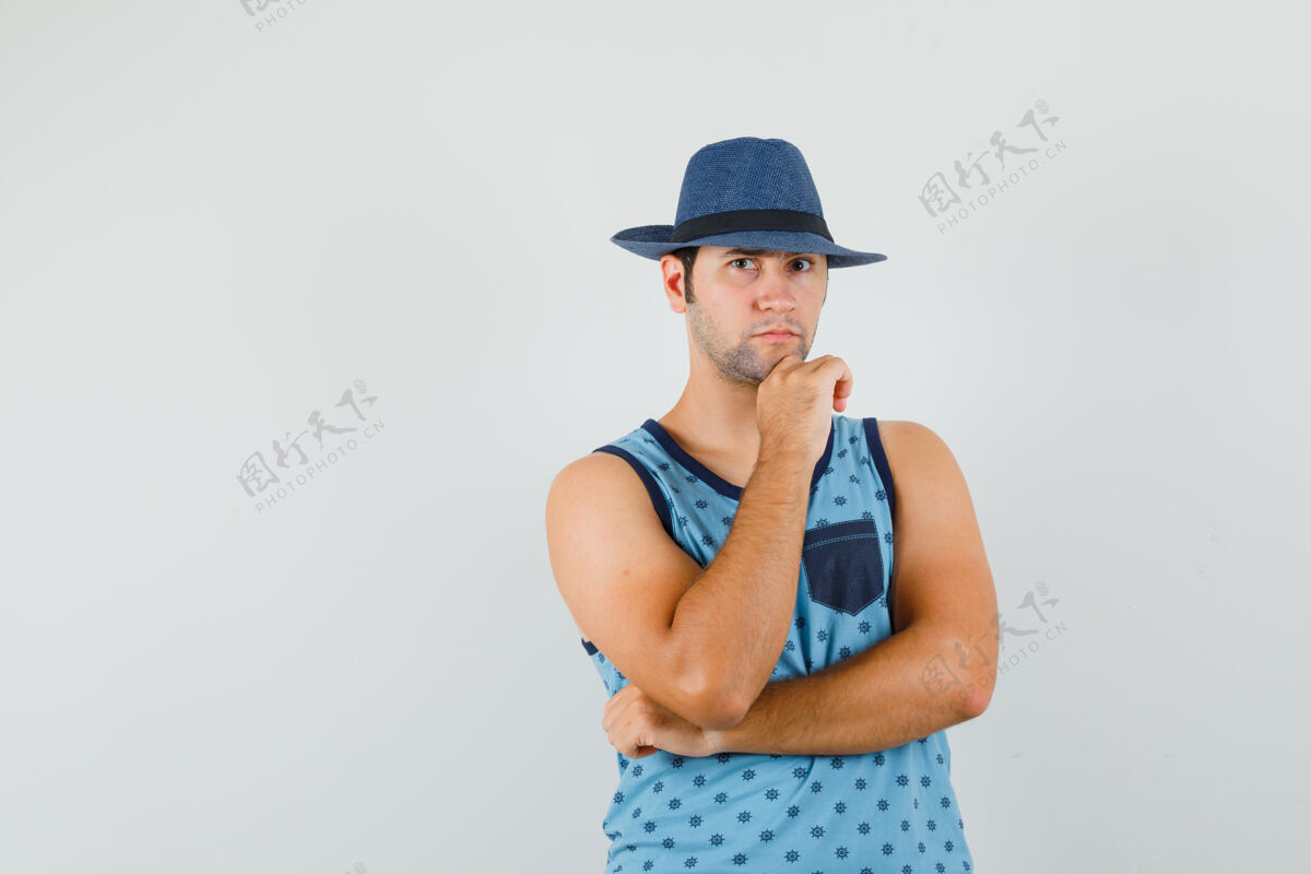 胡茬年轻人站在思考的姿势在蓝色的单打 帽子 并期待认真前视图发型微笑自信