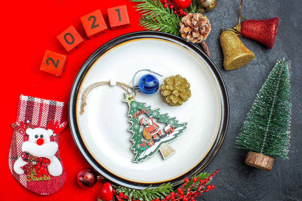 容器上图为新年背景 配餐盘装饰配件杉木树枝和数字圣诞袜 放在黑色桌子上圣诞树旁的红色餐巾上风景冷杉袜子