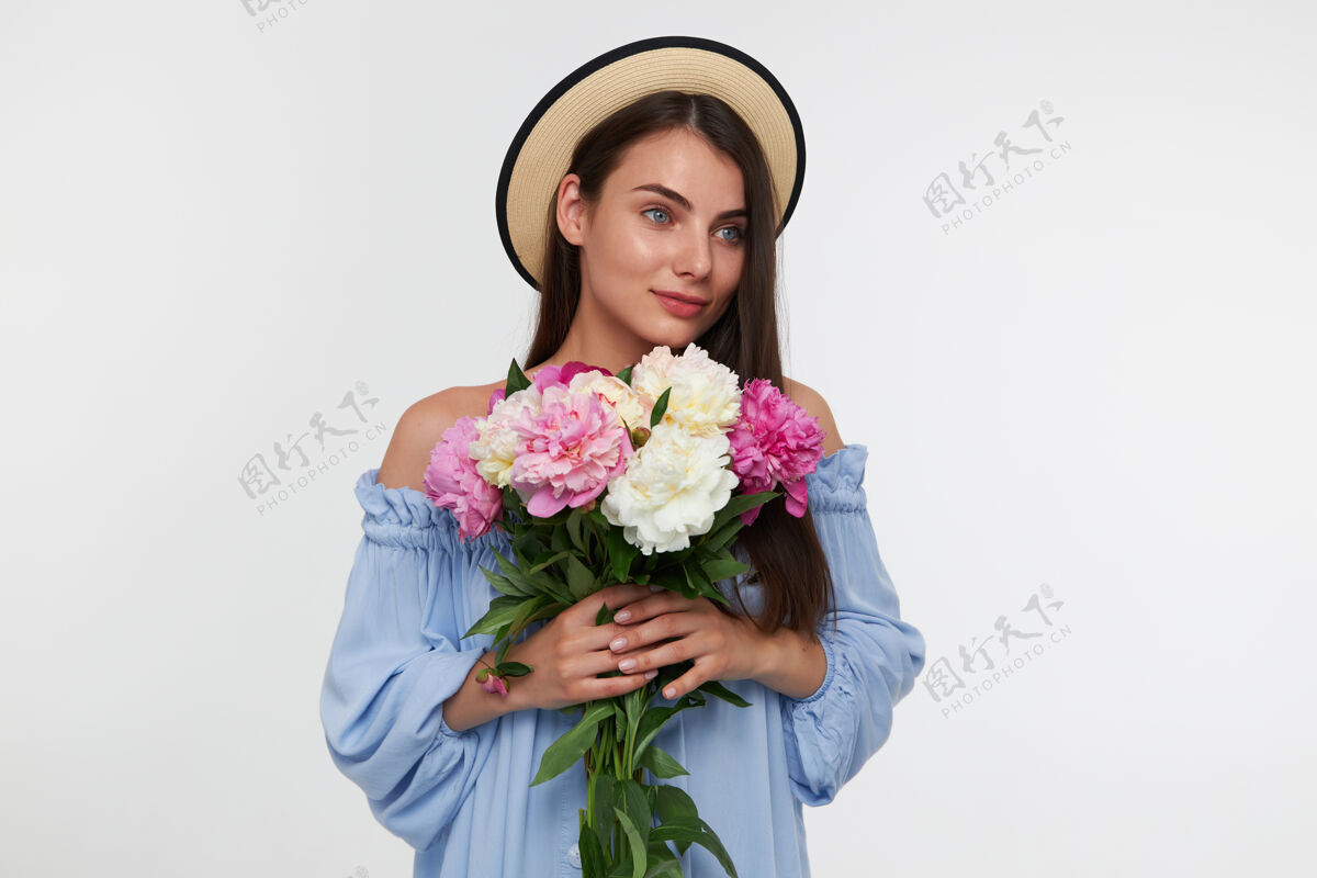 梦想年轻的女士 漂亮的女人 留着深褐色的长发戴着帽子 穿着蓝色漂亮的裙子手里拿着一束漂亮的花在白色墙壁上的复制空间向右看休闲面部花
