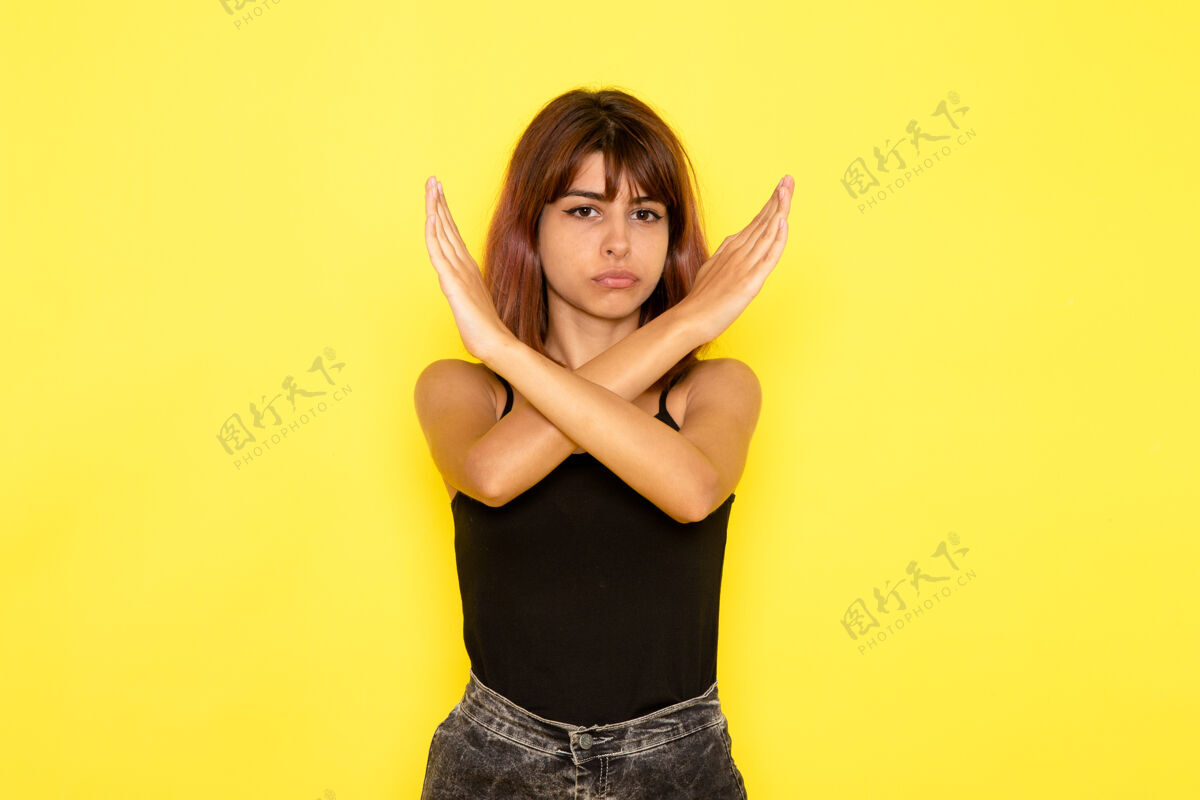 年轻身穿黑色衬衫和灰色牛仔裤的年轻女性正面图 黄色墙上显示禁止标志女性牛仔裤姿势