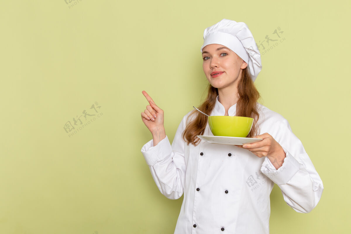 微笑身穿白色厨师服的女厨师手持浅绿色墙壁上的绿色盘子的正视图烹饪烹饪专业