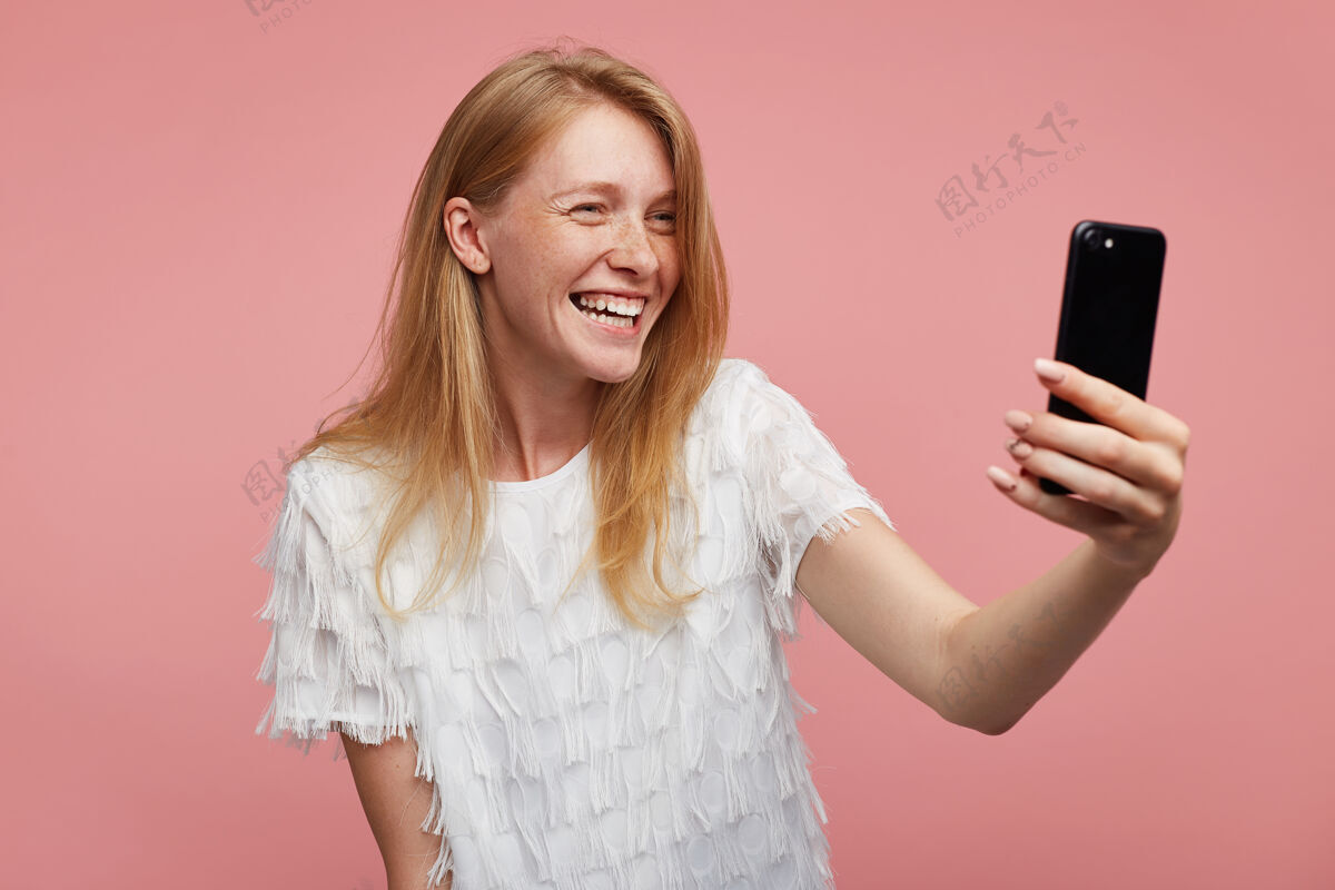 手快乐年轻可爱的红发女性 随意的发型 站在粉色的背景下 愉快地看着智能手机的镜头 微笑着自拍手机T恤姿势