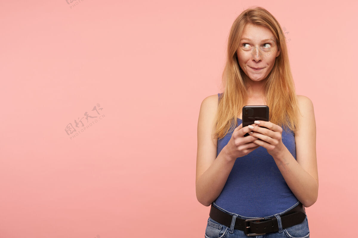 衬衫积极向上的年轻可爱的长发红发女士 穿着蓝色衬衫 手里拿着手机 若有所思地向上看 隔离在粉色背景下粉色手姿势